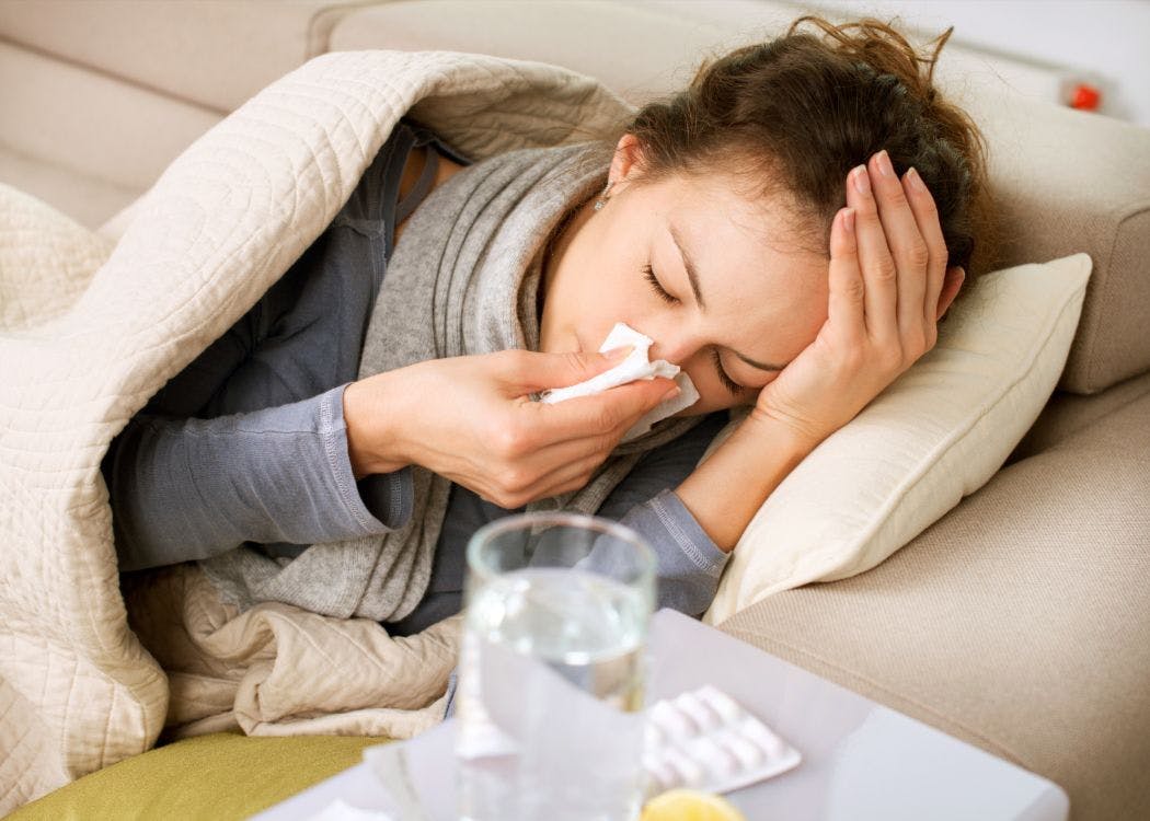 Sick woman wrapped in blankets blowing her nose with a tissue and holding head